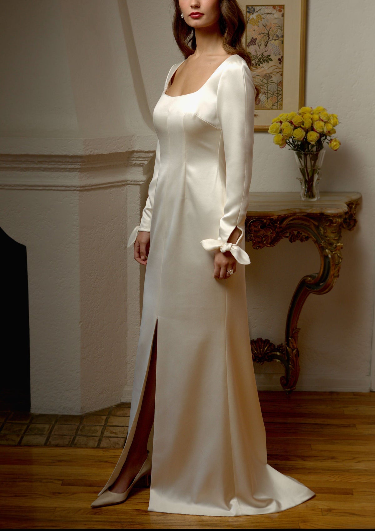 Three-quarter view of a fitted Créme satin gown with long sleeves, keyhole wrist ties, and a train pooling on the floor