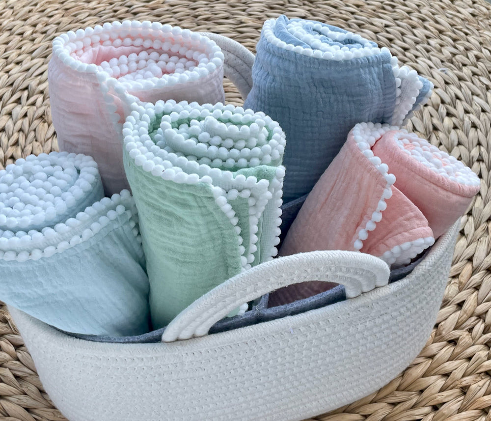Top view of cotton muslin swaddle blankets showcasing pastel colors and pom-pom trim in a white basket on a woven mat