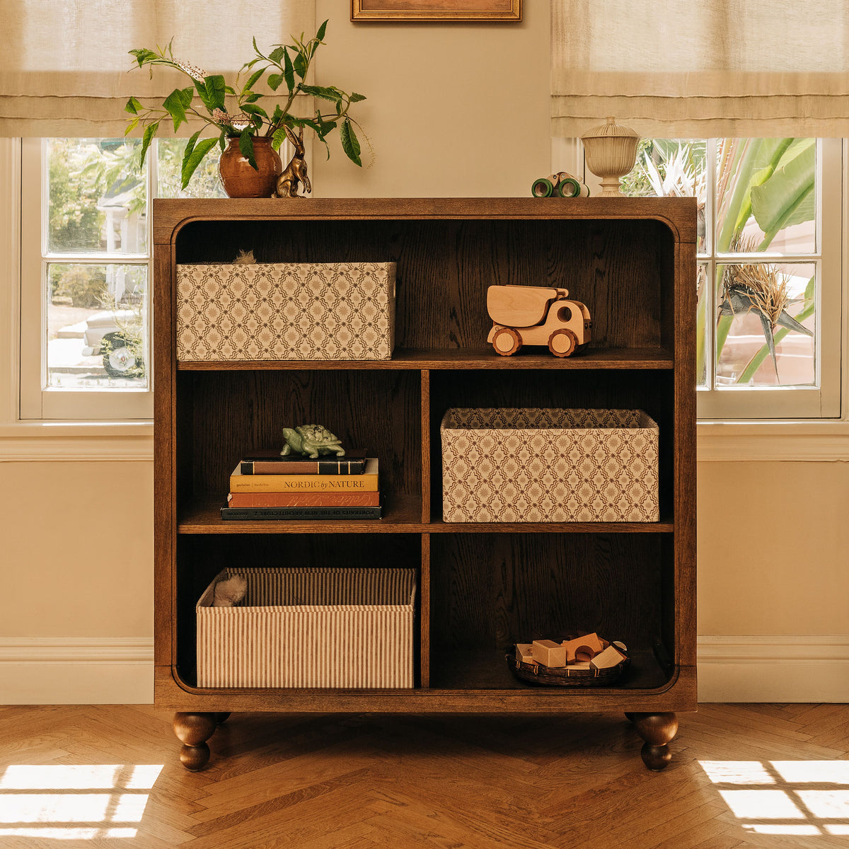 Front view of Astre Assembled Bookcase by Nate Berkus in Toffee with six compartments and rounded feet in a sunlit room