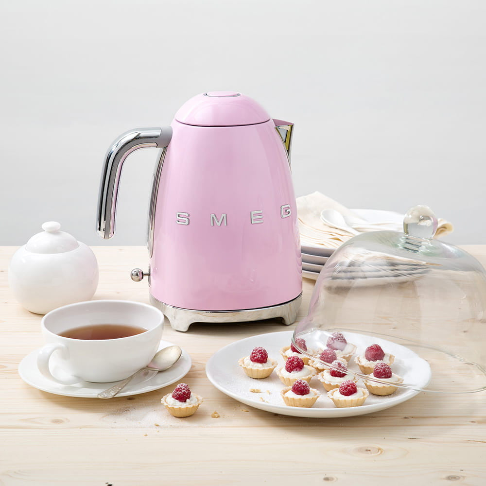 Angled view of pink SMEG kettle with chrome handle and logo on light wood table beside teacup and plate of raspberry tartlets