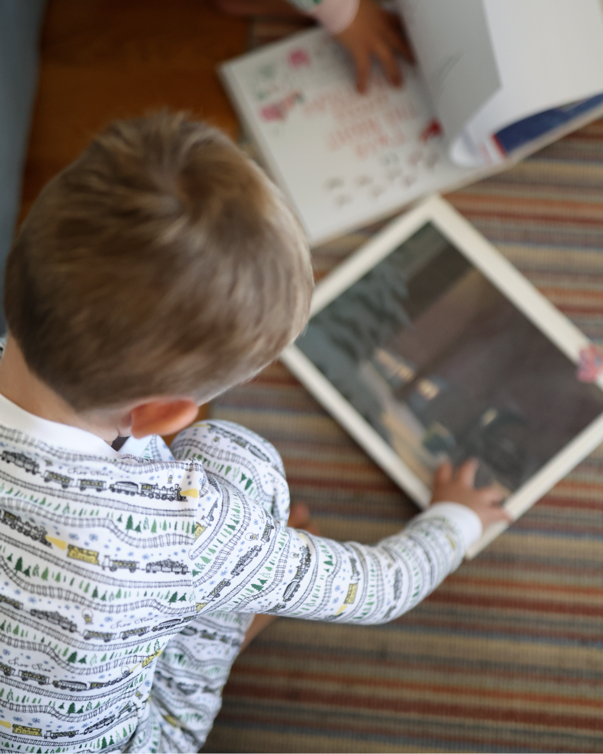 Overhead view of Christmas Trains Kids Pajamas, Set of 2 showcasing the train print on a child using a tablet beside a book