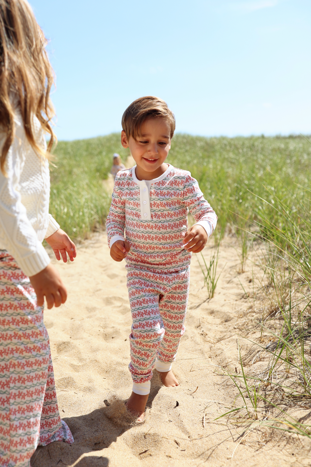 Child wearing Christmas lobster henley pajamas walking on a sandy beach with another child nearby