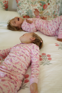 Top view of two children wearing matching pink unicorn two-piece kids pajamas, showcasing the patterned print on a floral bed