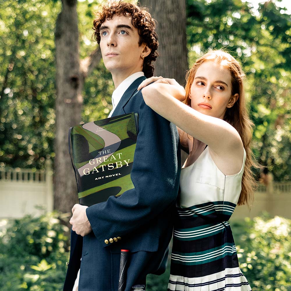 Young man holding The Great Gatsby art novel while a young woman leans on his shoulder in a sunlit garden
