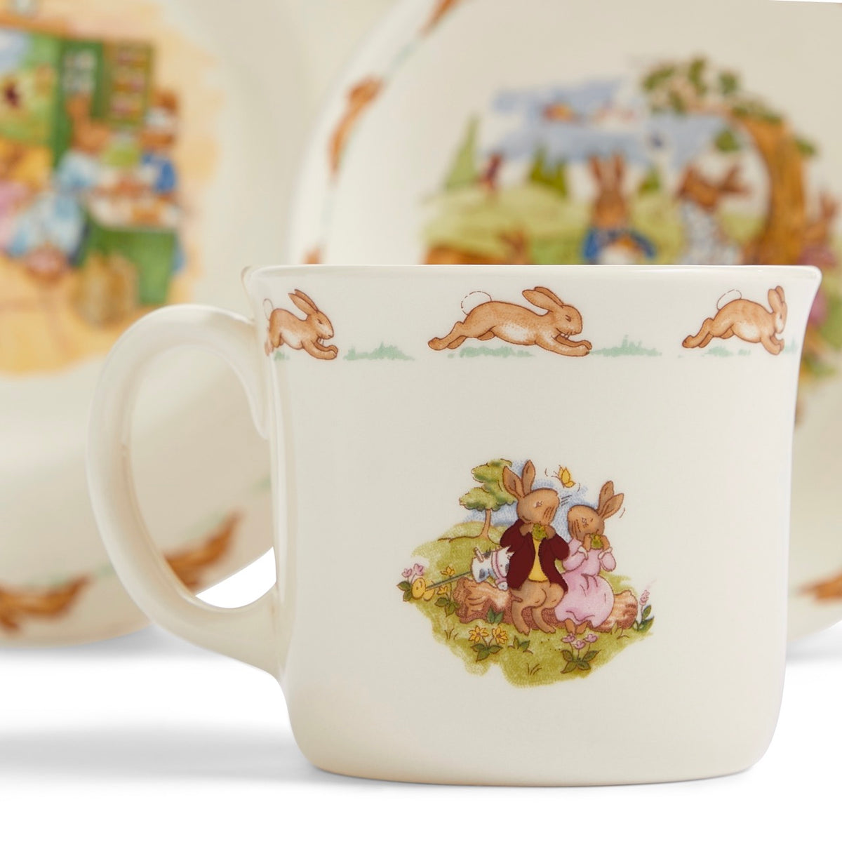 Close-up of Bunnykins Childrens Bowl, Plate and Mug, three-piece set showcasing rabbit illustrations with blurred dishes behind