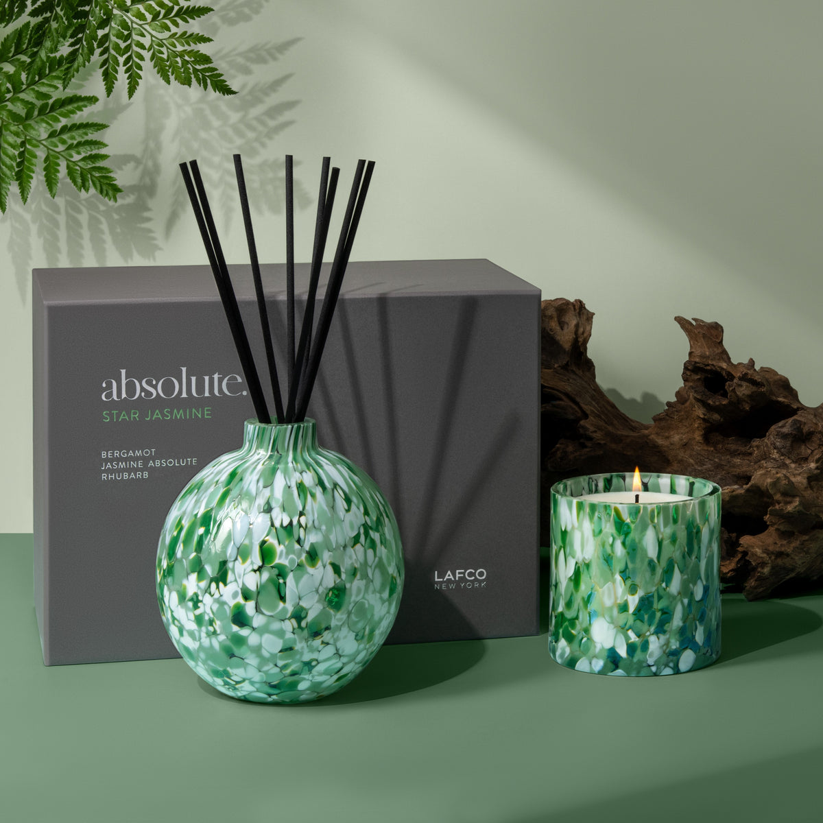 Close up of Star Jasmine Absolute diffuser with black reeds and lit matching candle in green speckled glass beside gray box