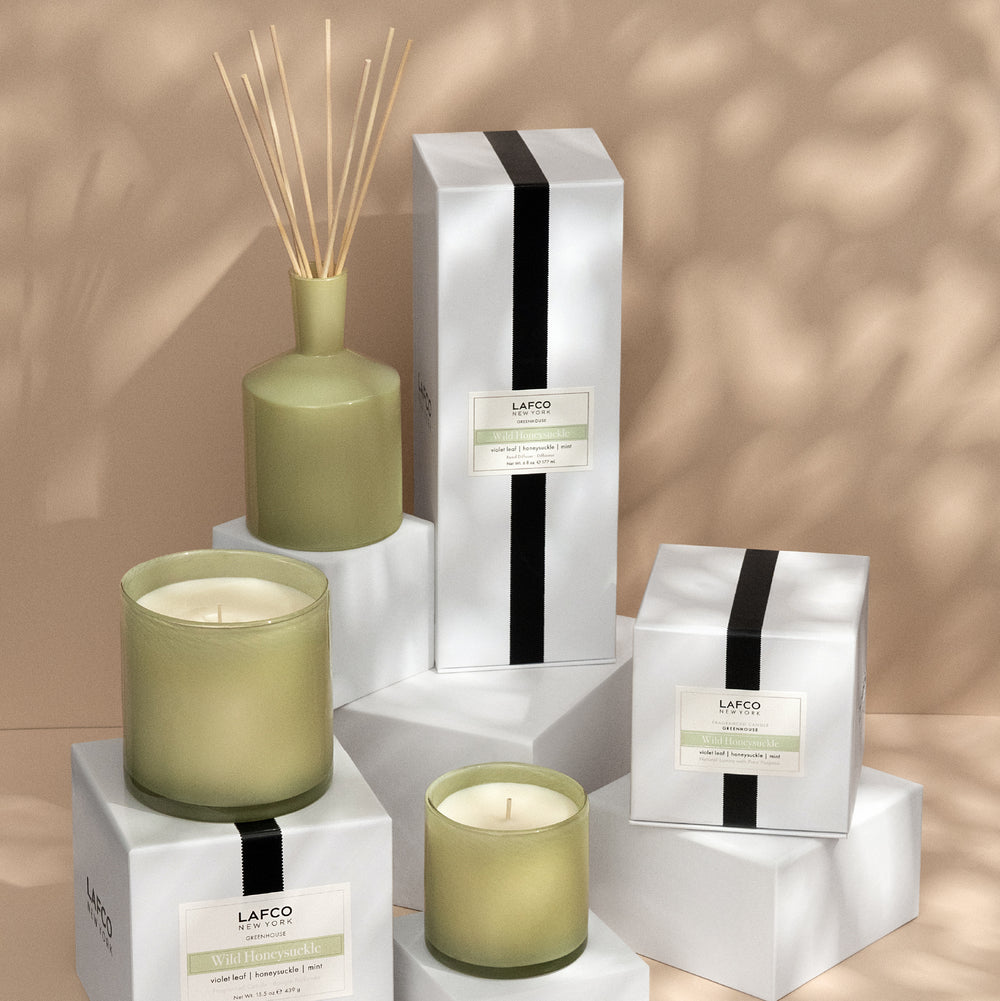 Front view of Wild Honeysuckle classic candle and reed diffuser in green glass on white display boxes against a warm beige background