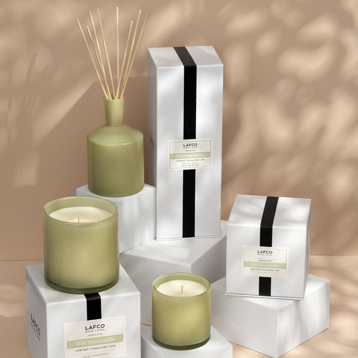 Front view of Wild Honeysuckle classic candle and reed diffuser in green glass on white display boxes against a warm beige background