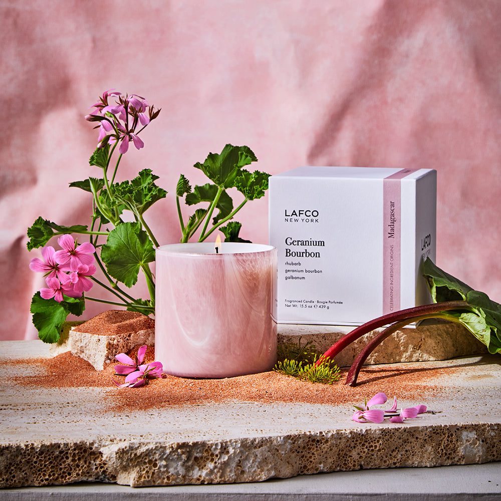 Front view of Geranium Bourbon scented candle in pink marbled glass, lit beside branded box and geranium flowers on stone slab