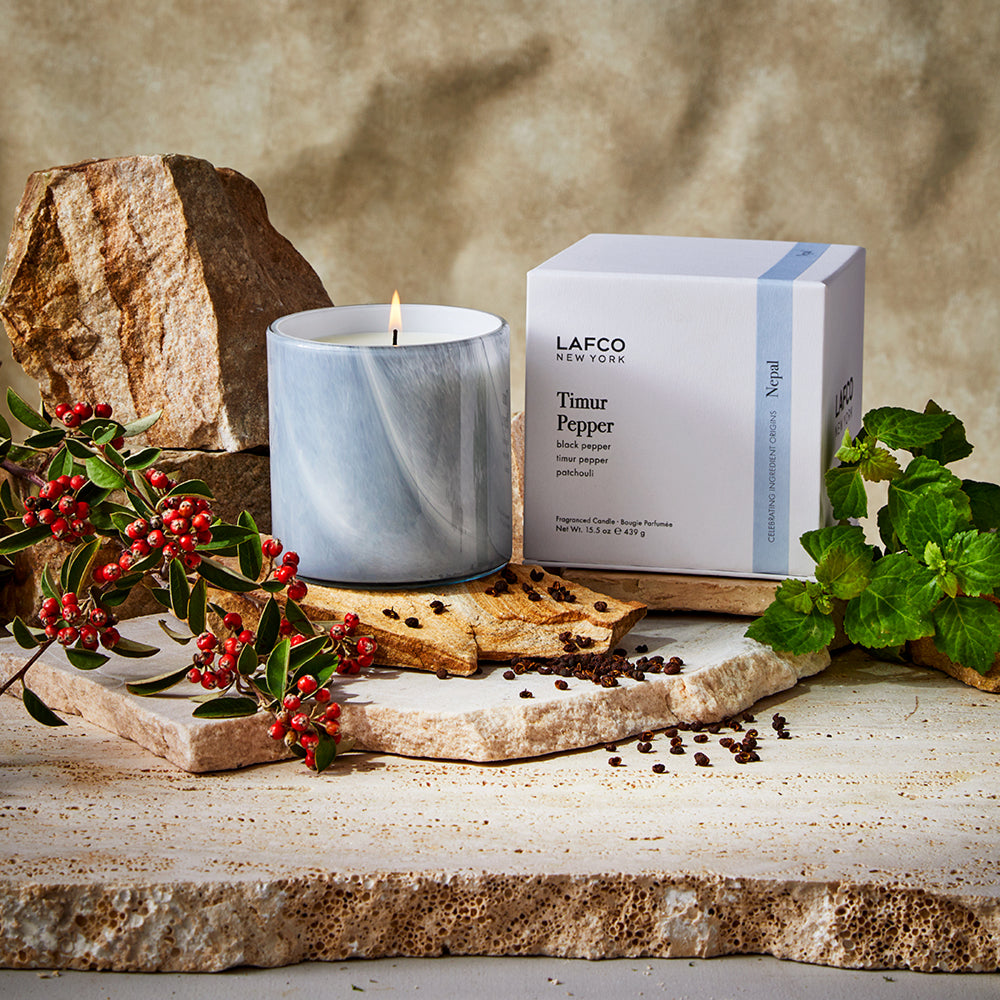Front view of Timur Pepper signature candle in blue-gray marble jar, lit, with labeled box, scattered peppercorns and red berries on a stone slab background