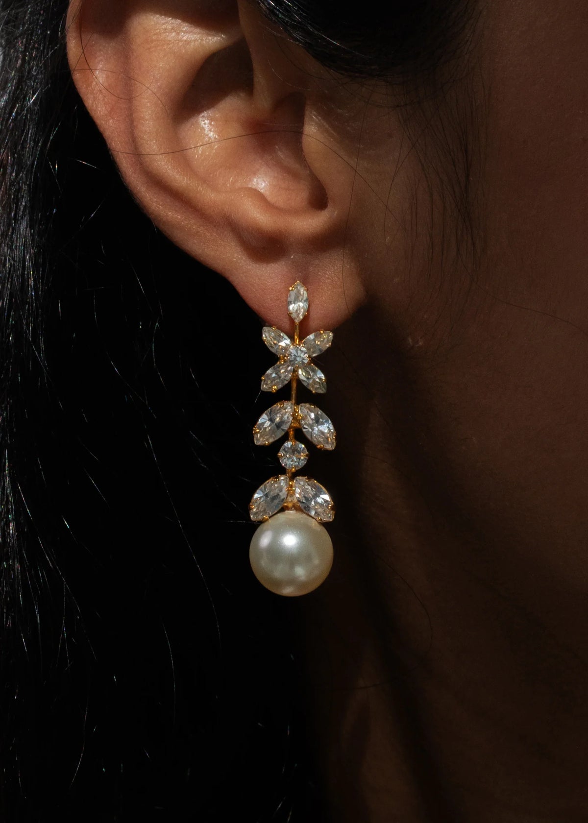 Close-up of Tiera earring in crystal antique gold showing linear Austrian crystals and a pearl drop on a model’s ear against a dark background