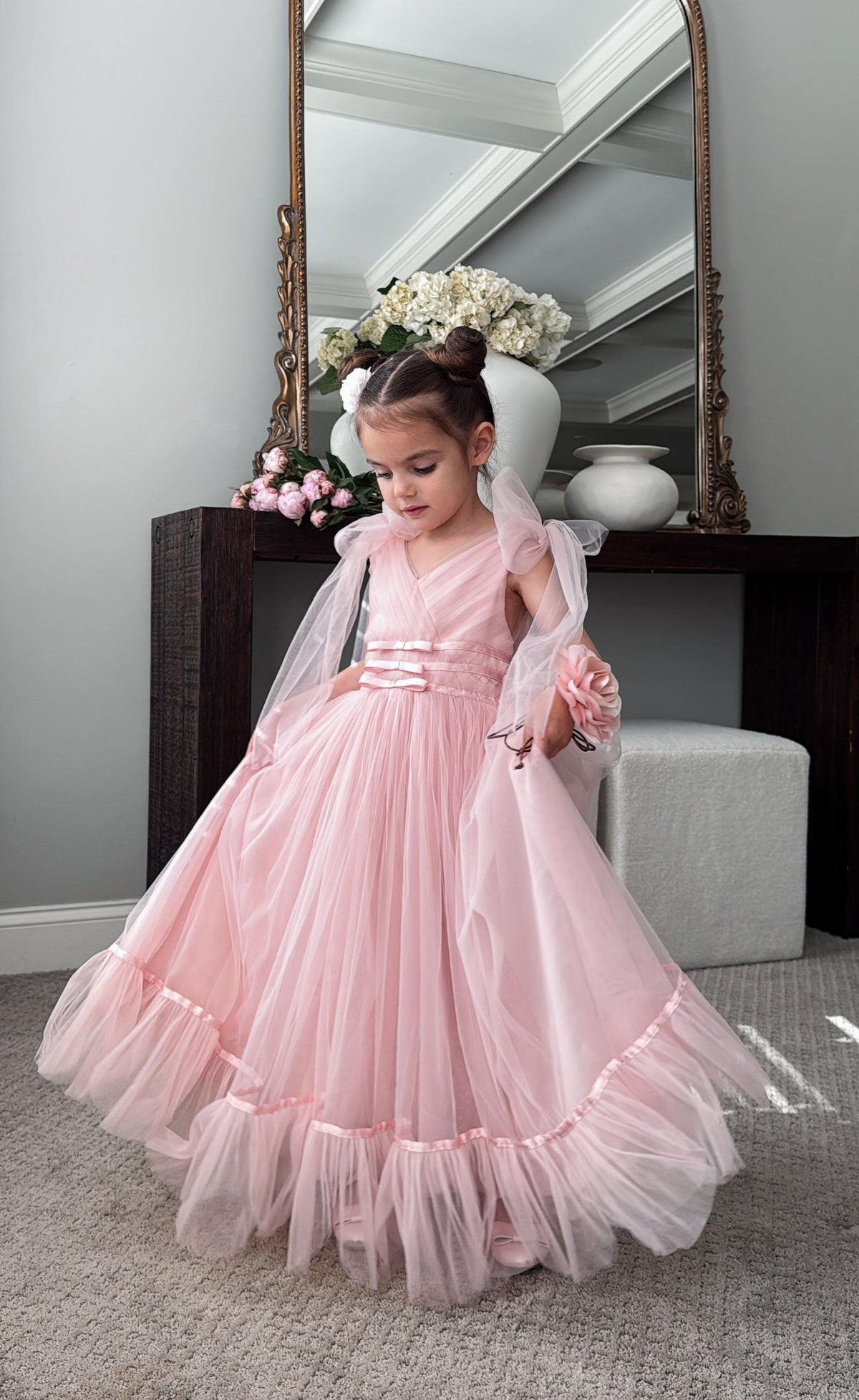Light pink tulle flower girl dress with V-neck bodice and soft tiered skirt, styled indoors by mirror.