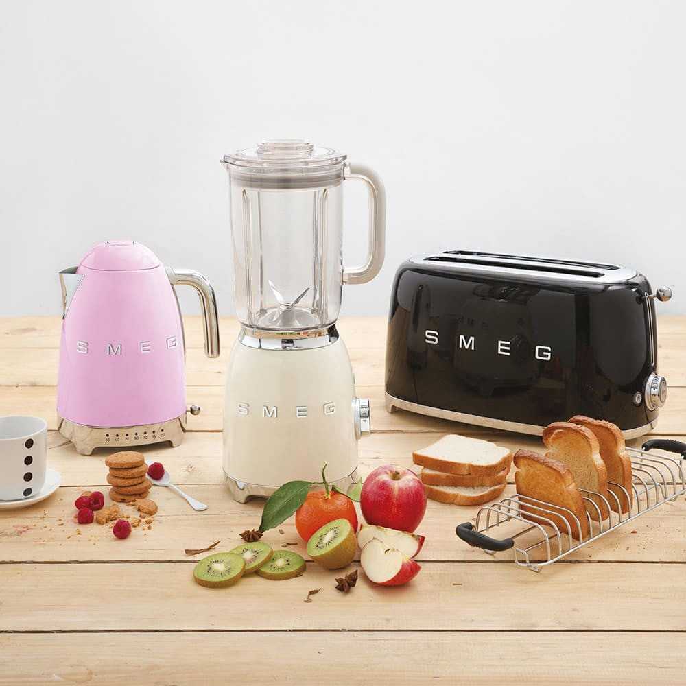Pink SMEG kettle with cream blender and black toaster on wooden table with fruit and toast, breakfast scene