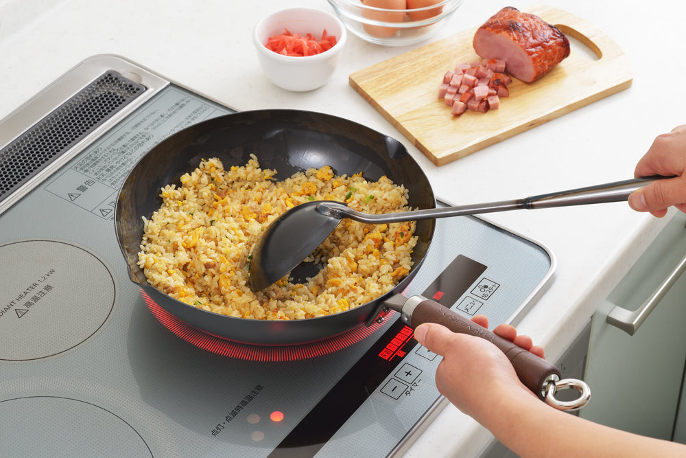 Black carbon steel deep fry pan cooking fried rice on an induction cooktop, hand holding the handle and a spatula