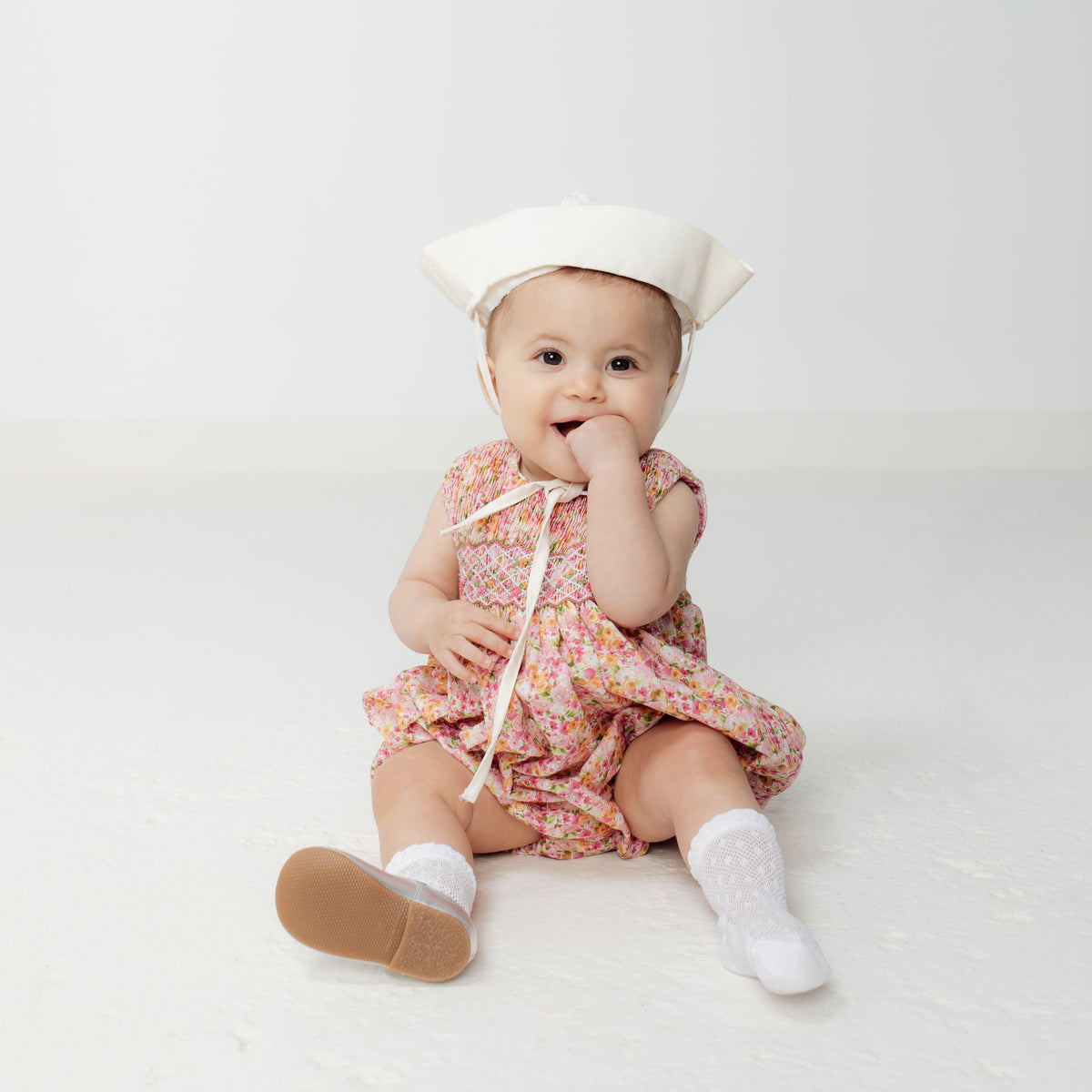 Seated view of Isabel Bubble romper on baby model showing pink floral linen, smocked front, and snap bottom on white background