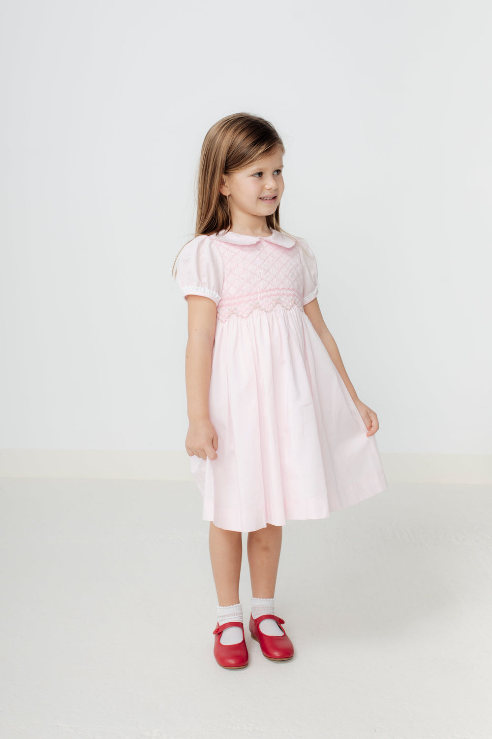 Front view of Sofia Dress in pale pink on child model showing smocked panel, puff sleeves, Peter Pan collar against white