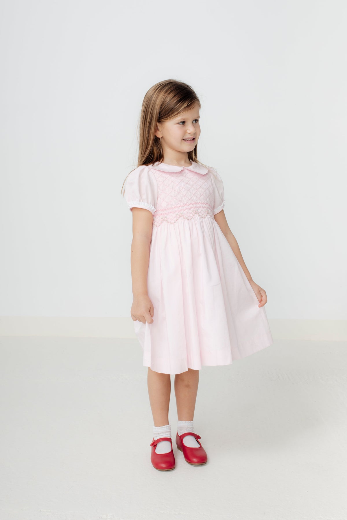 Front view of Sofia Dress in pale pink on child model showing smocked panel, puff sleeves, Peter Pan collar against white
