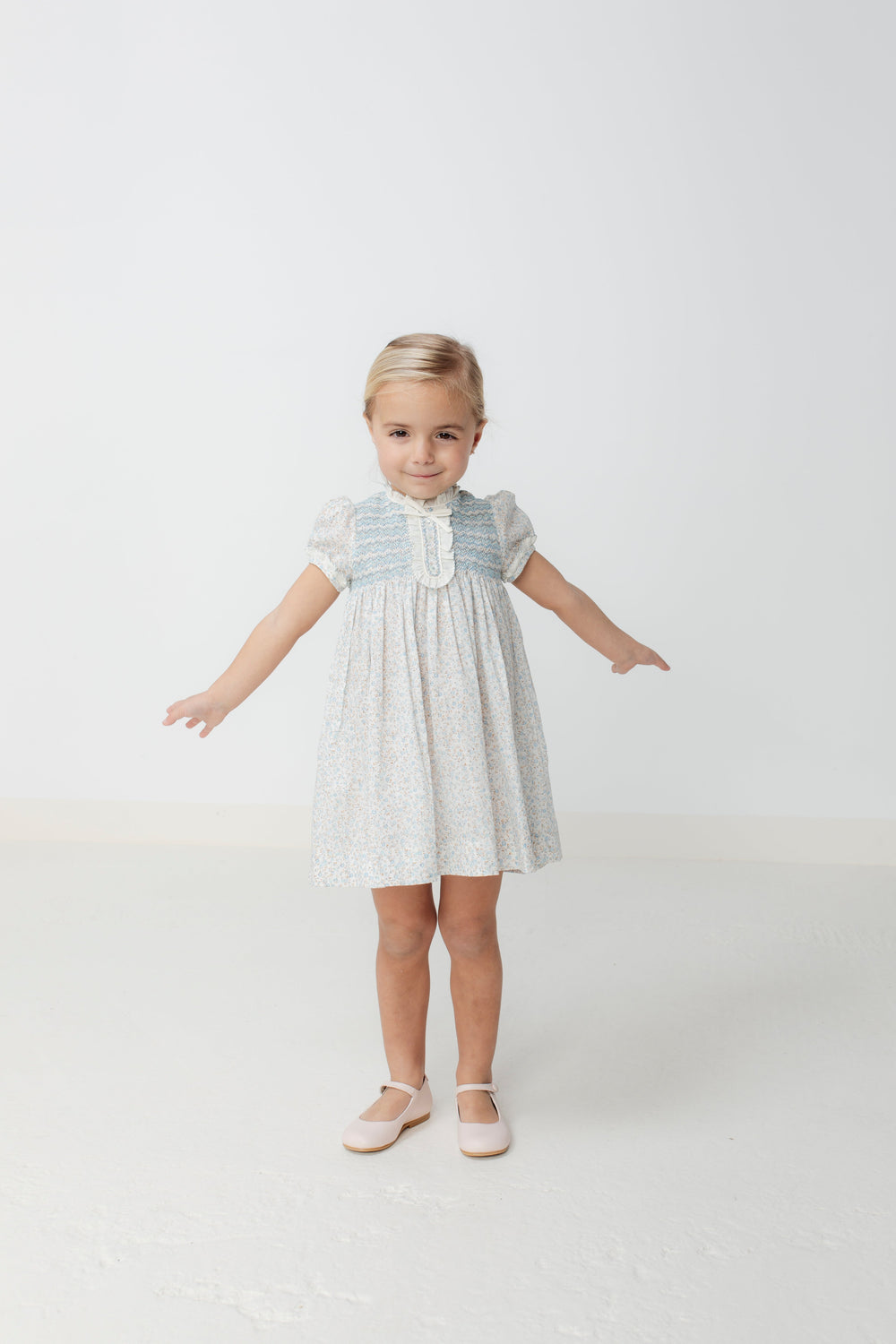 Front view of Clotilde Dress in blue on child model, smocked bodice, puff sleeves, knee-length floral skirt