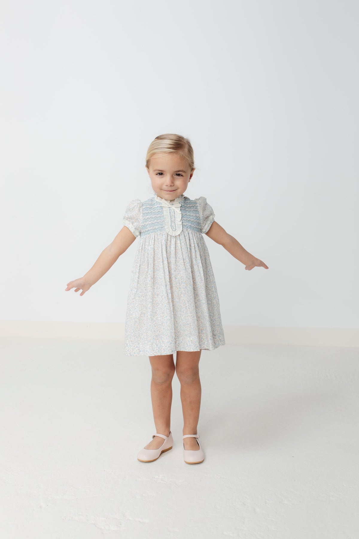 Front view of Clotilde Dress in blue on child model, smocked bodice, puff sleeves, knee-length floral skirt