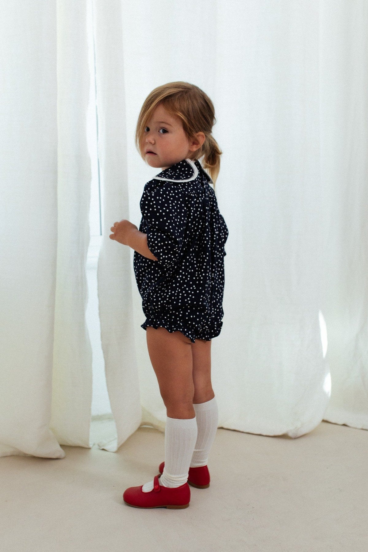 Side view of Clementine Bubble in Polka Dots with oversized white petal collar, worn by a child before white curtains