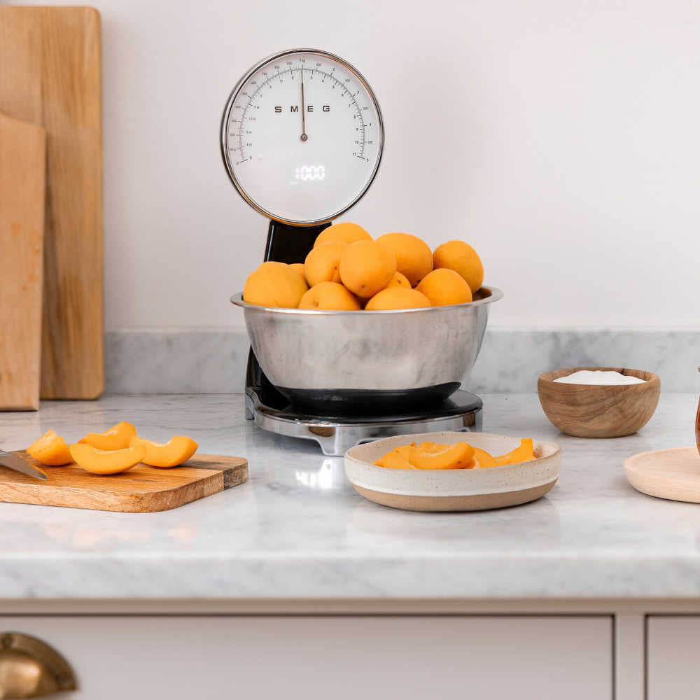 SMEG 50s kitchen scale with stainless-steel bowl of apricots on a marble countertop and sliced apricot on a cutting board