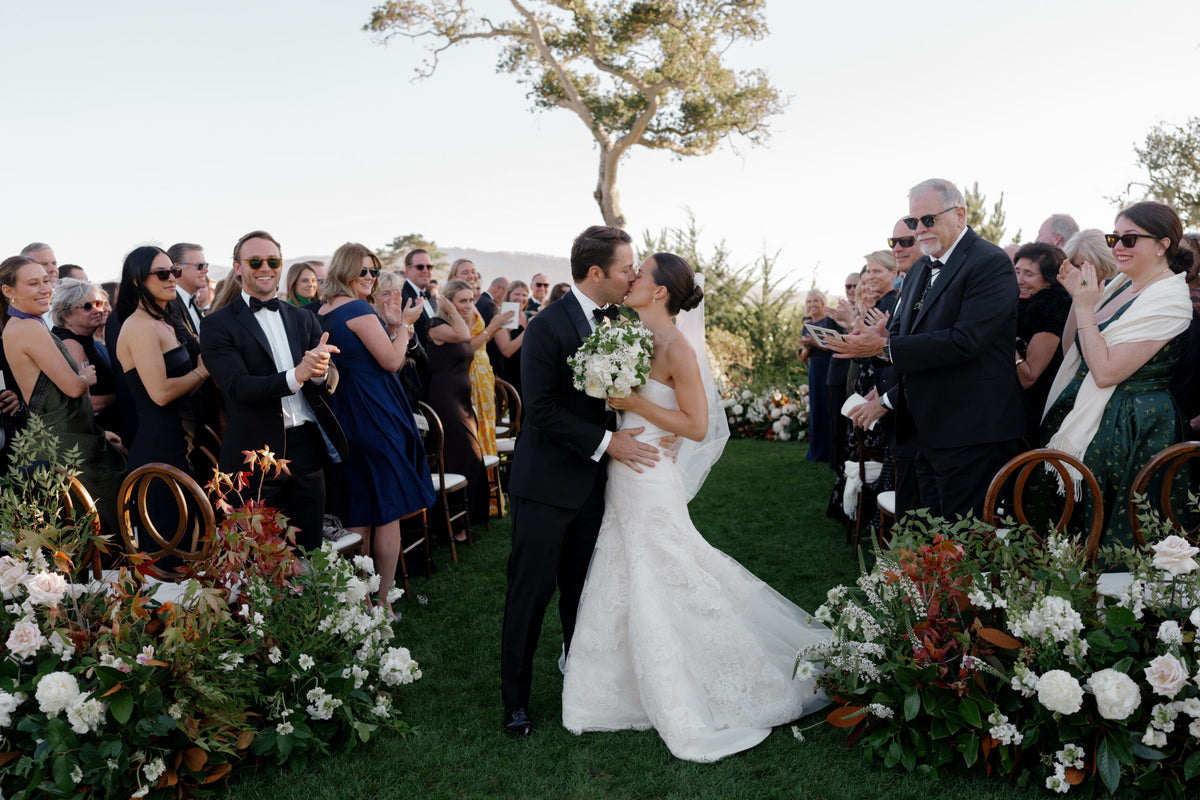 Bride and groom kiss during their Pebble Beach wedding ceremony surrounded by guests and flowers