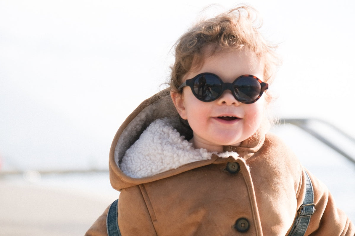 Toddler smiling wearing tortoise round Joy infant sunglasses outdoors in a hooded shearling-lined coat