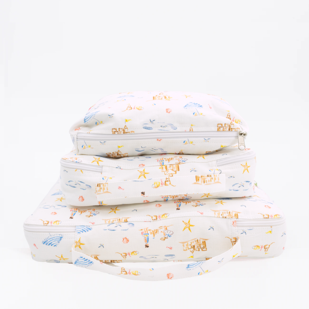 Front view of Sandy packing cubes, three stacked, showing beach-scene print, zipper closures and fabric handle on white background