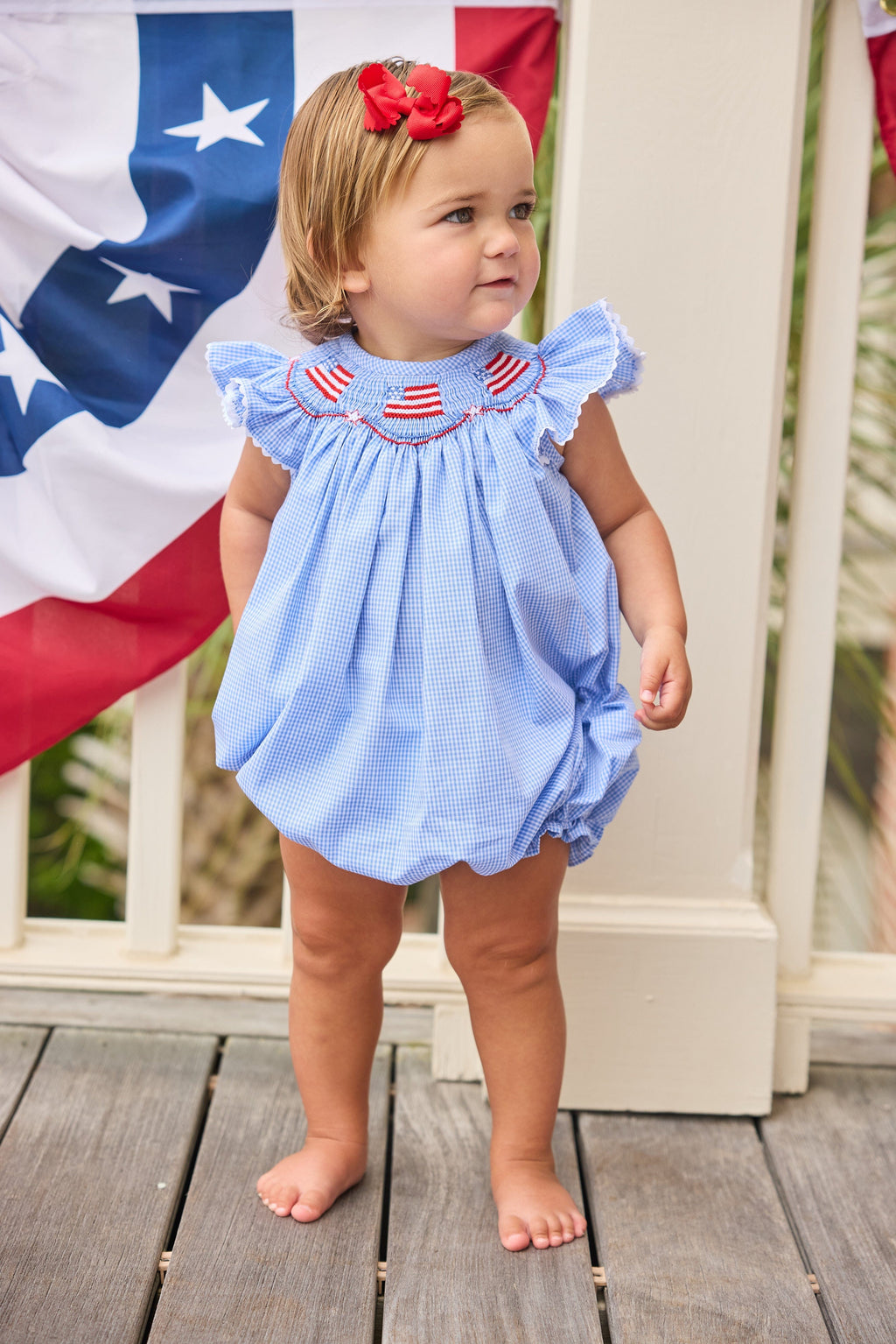 Little English traditional children's clothing.  Flutter sleeve bubble in royal blue mini check fabric and smocking with American flag bunting for baby girls