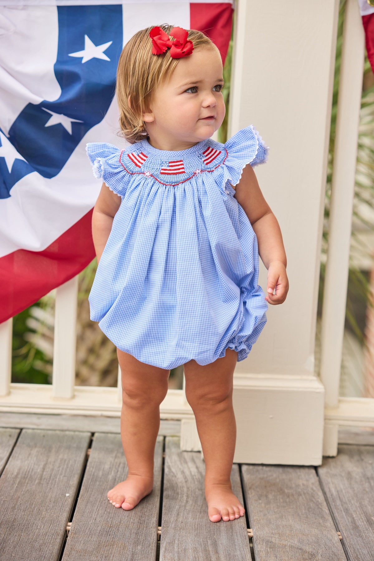 Little English traditional children's clothing.  Flutter sleeve bubble in royal blue mini check fabric and smocking with American flag bunting for baby girls