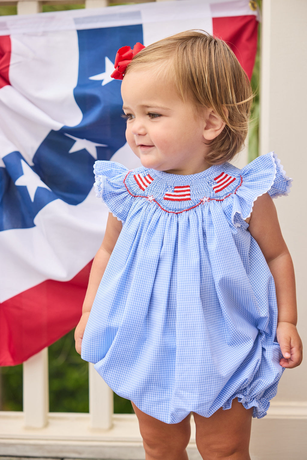 Little English traditional children's clothing.  Flutter sleeve bubble in royal blue mini check fabric and smocking with American flag bunting for baby girls