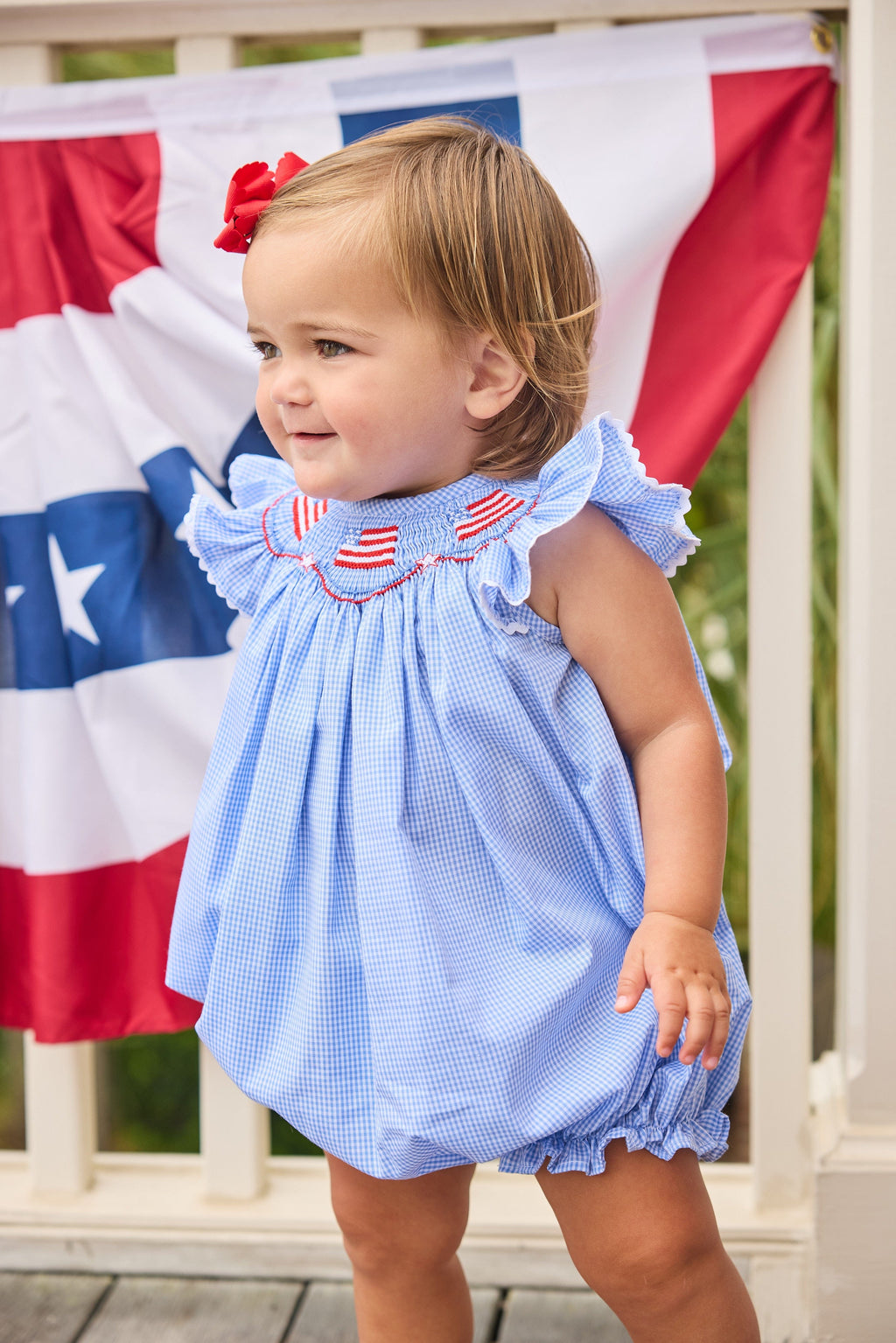 Little English traditional children's clothing.  Flutter sleeve bubble in royal blue mini check fabric and smocking with American flag bunting for baby girls