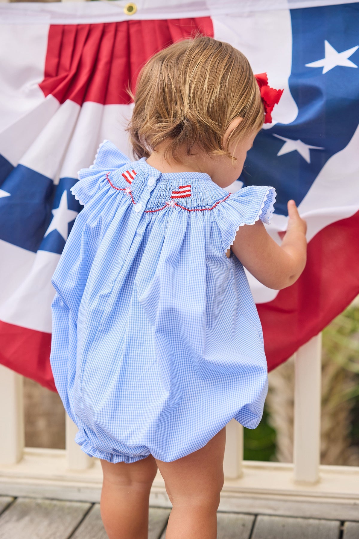 Little English traditional children's clothing.  Flutter sleeve bubble in royal blue mini check fabric and smocking with American flag bunting for baby girls