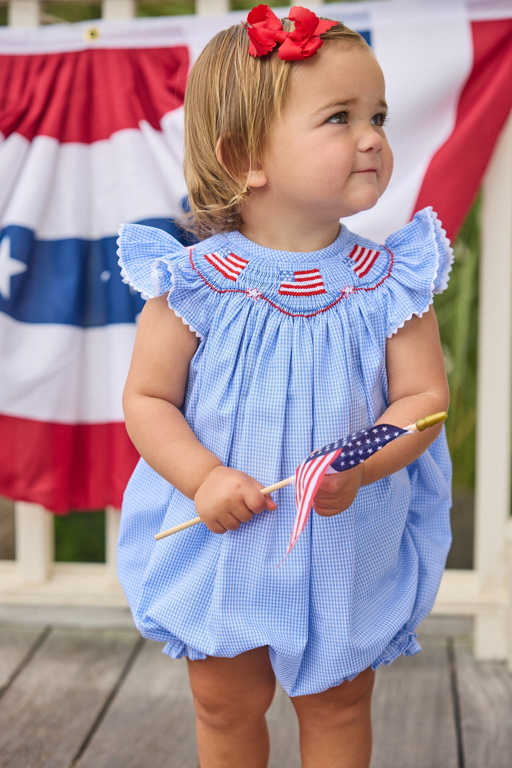 Little English traditional children's clothing.  Flutter sleeve bubble in royal blue mini check fabric and smocking with American flag bunting for baby girls