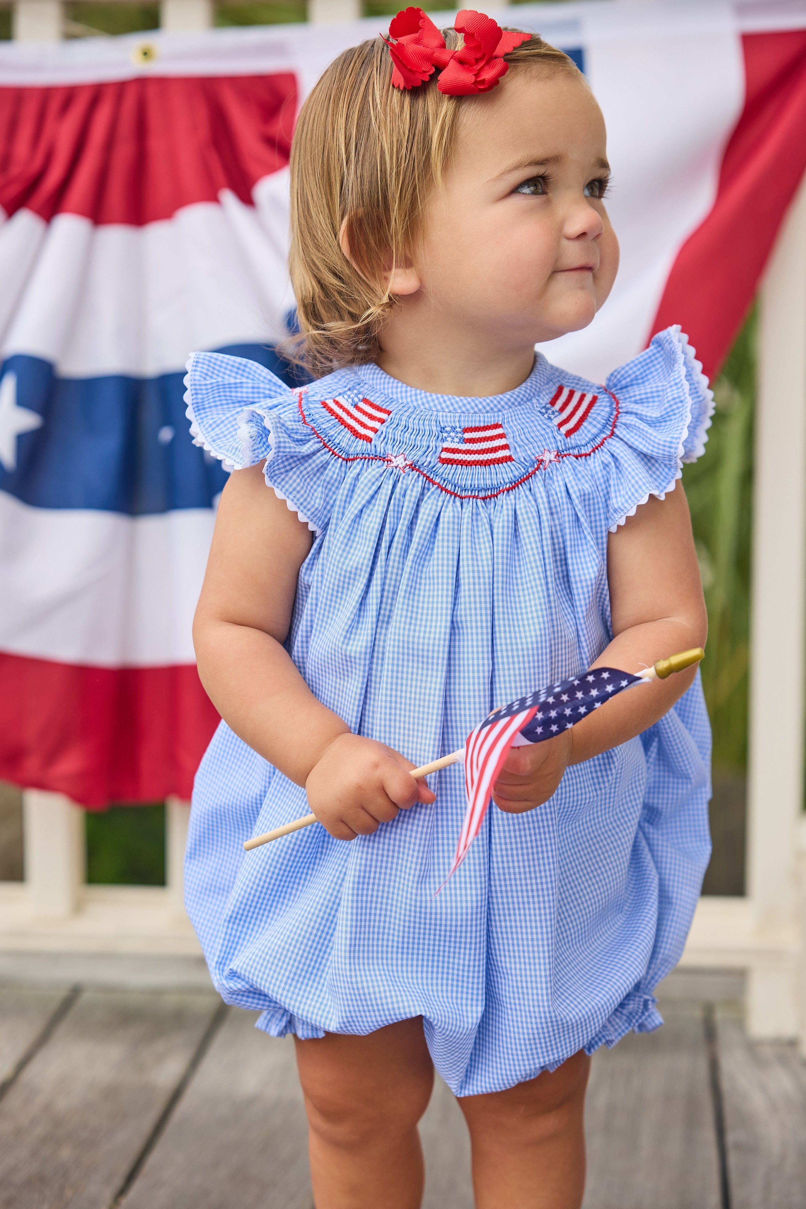 Little English traditional children's clothing.  Flutter sleeve bubble in royal blue mini check fabric and smocking with American flag bunting for baby girls