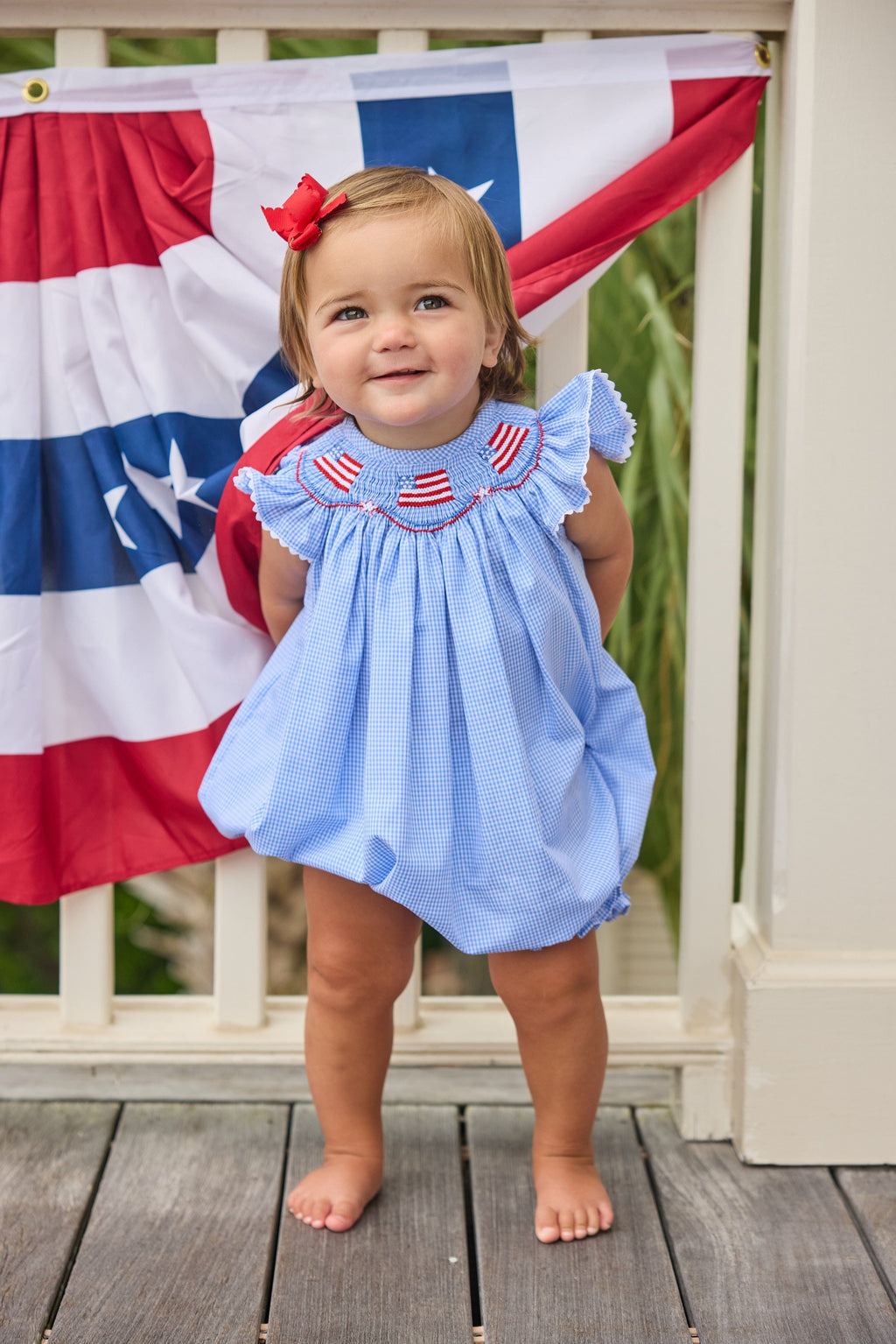 Little English traditional children's clothing.  Flutter sleeve bubble in royal blue mini check fabric and smocking with American flag bunting for baby girls