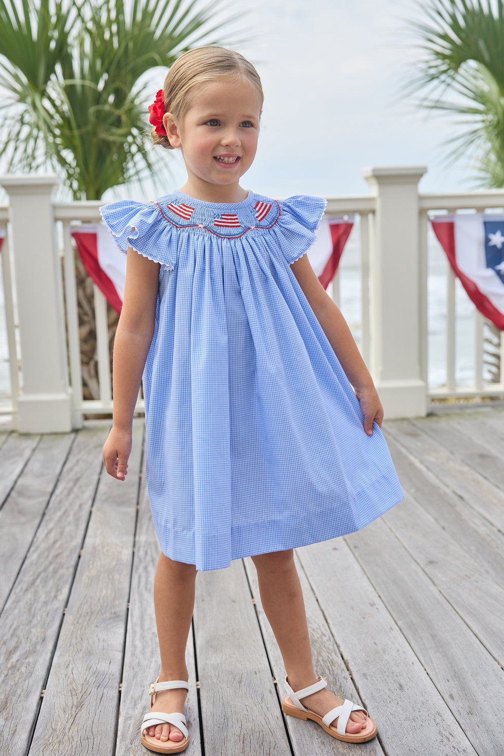 Little English traditional children's clothing. Flutter sleeve dress for toddler girls in royal blue mini gingham with smocking at the collar and American flag bunting