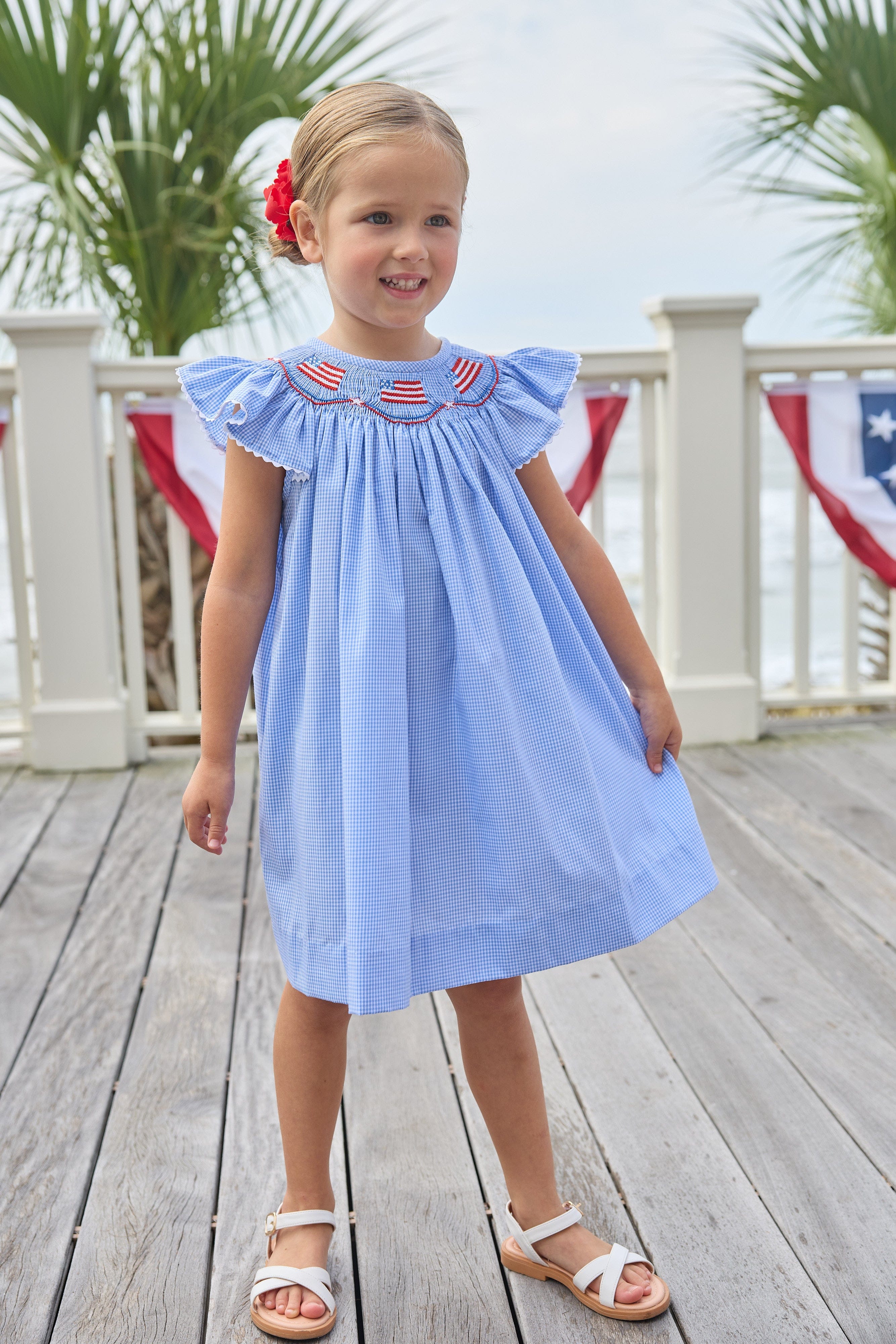 Little English traditional children's clothing. Flutter sleeve dress for toddler girls in royal blue mini gingham with smocking at the collar and American flag bunting