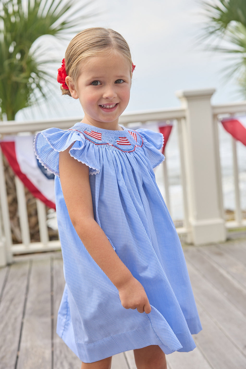Little English traditional children's clothing. Flutter sleeve dress for toddler girls in royal blue mini gingham with smocking at the collar and American flag bunting