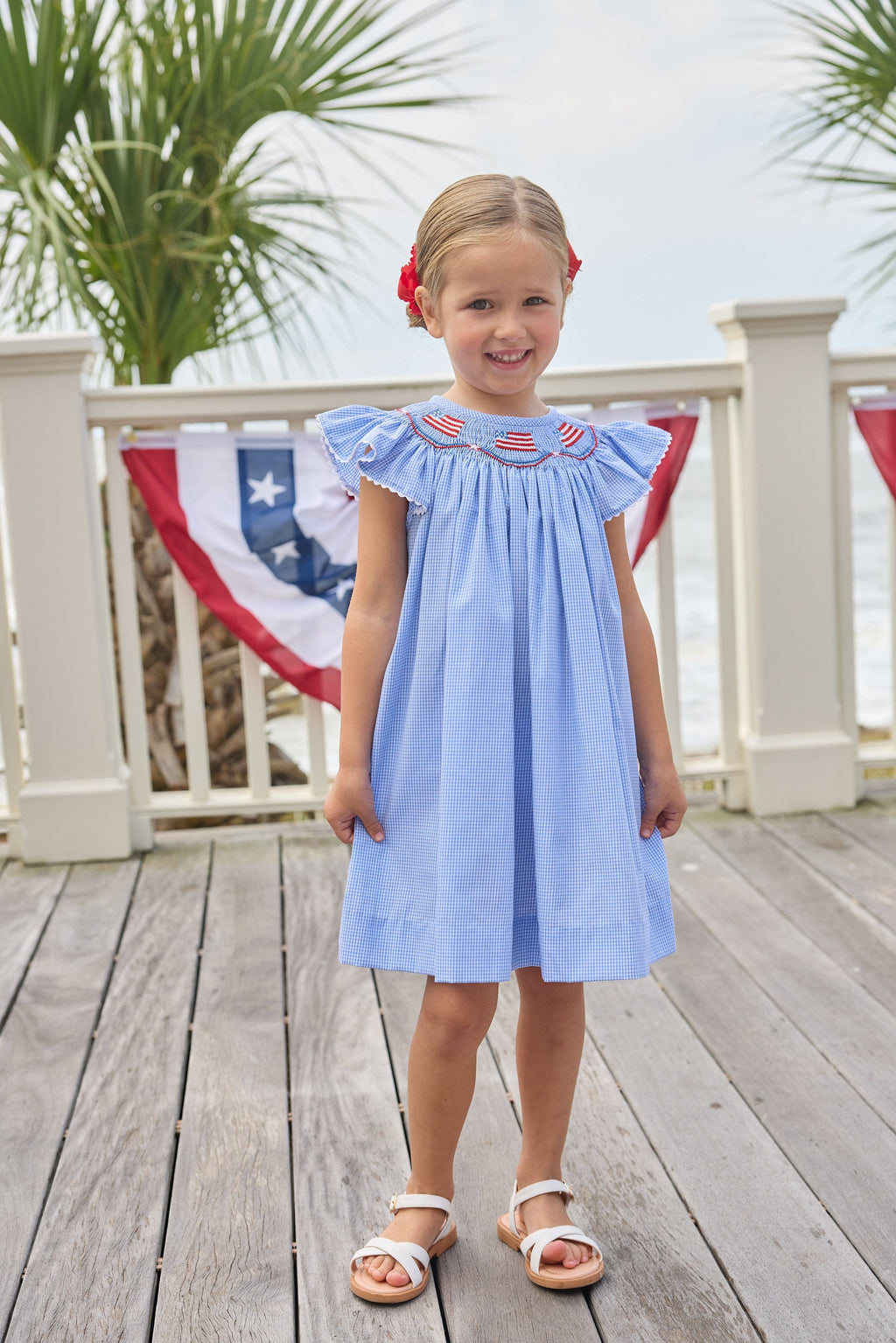 Little English traditional children's clothing. Flutter sleeve dress for toddler girls in royal blue mini gingham with smocking at the collar and American flag bunting