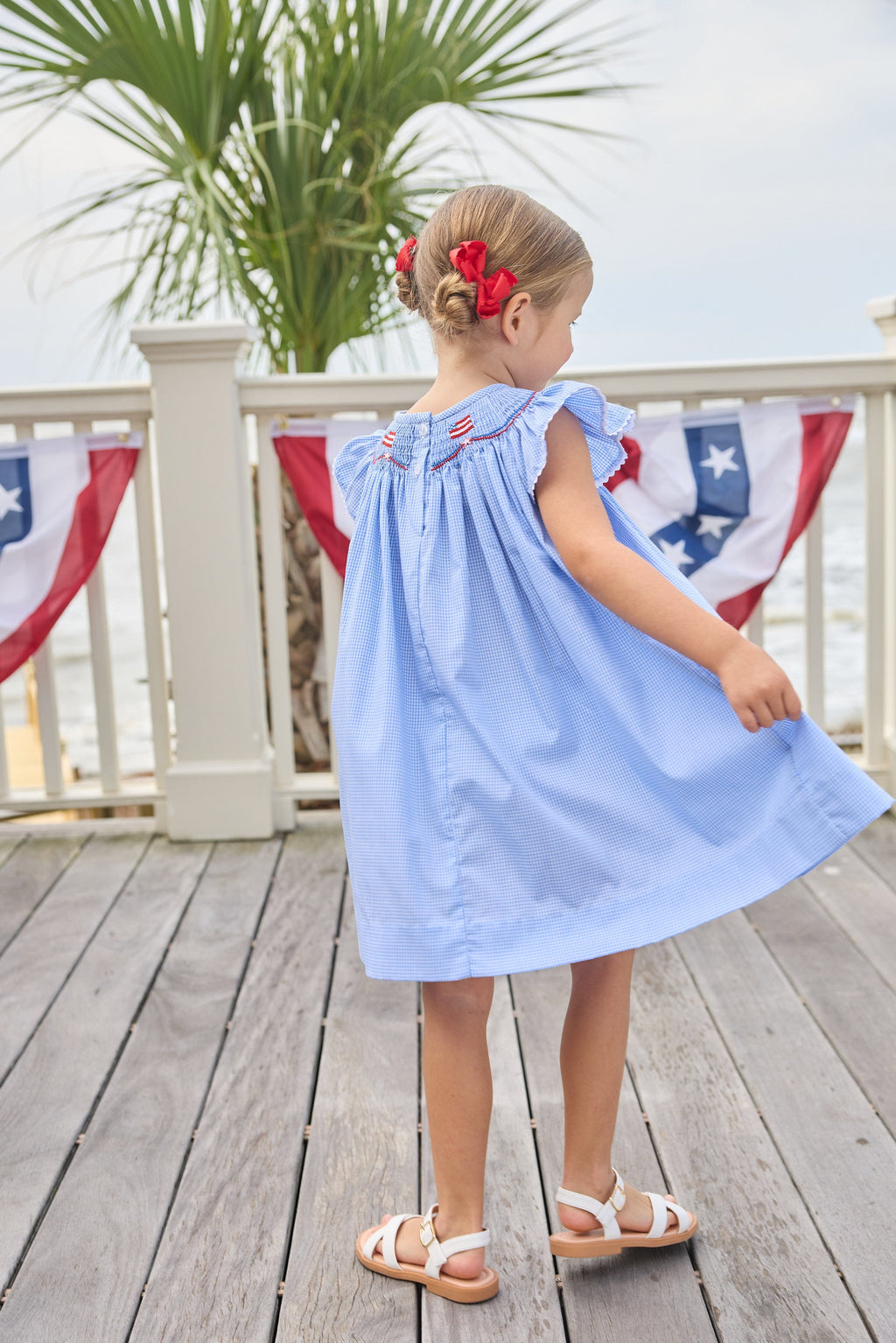 Little English traditional children's clothing. Flutter sleeve dress for toddler girls in royal blue mini gingham with smocking at the collar and American flag bunting