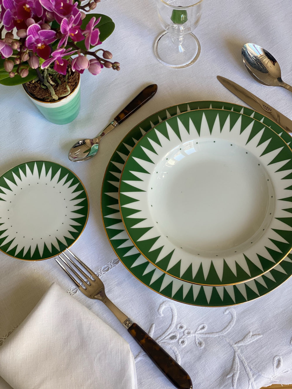Angled overhead view of Green Punk soup and pasta plate showcasing green starburst border and gold rim on a white embroidered tablecloth