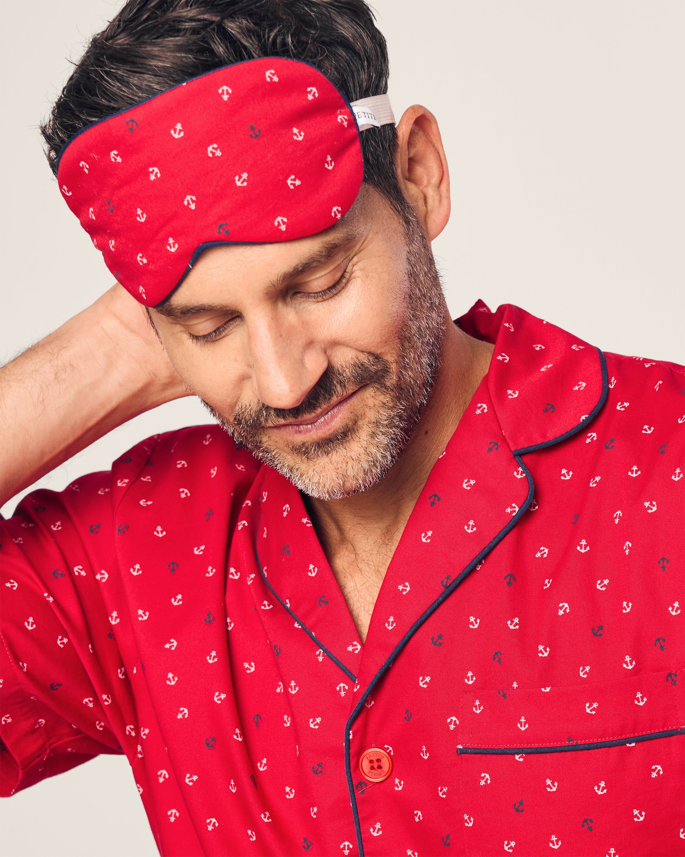 A person in red pajamas with a nautical print and wearing Petite Plumes Adults Twill Sleep Mask in Anchors Away scratches their neck, smiling contentedly. The background is a plain light color.