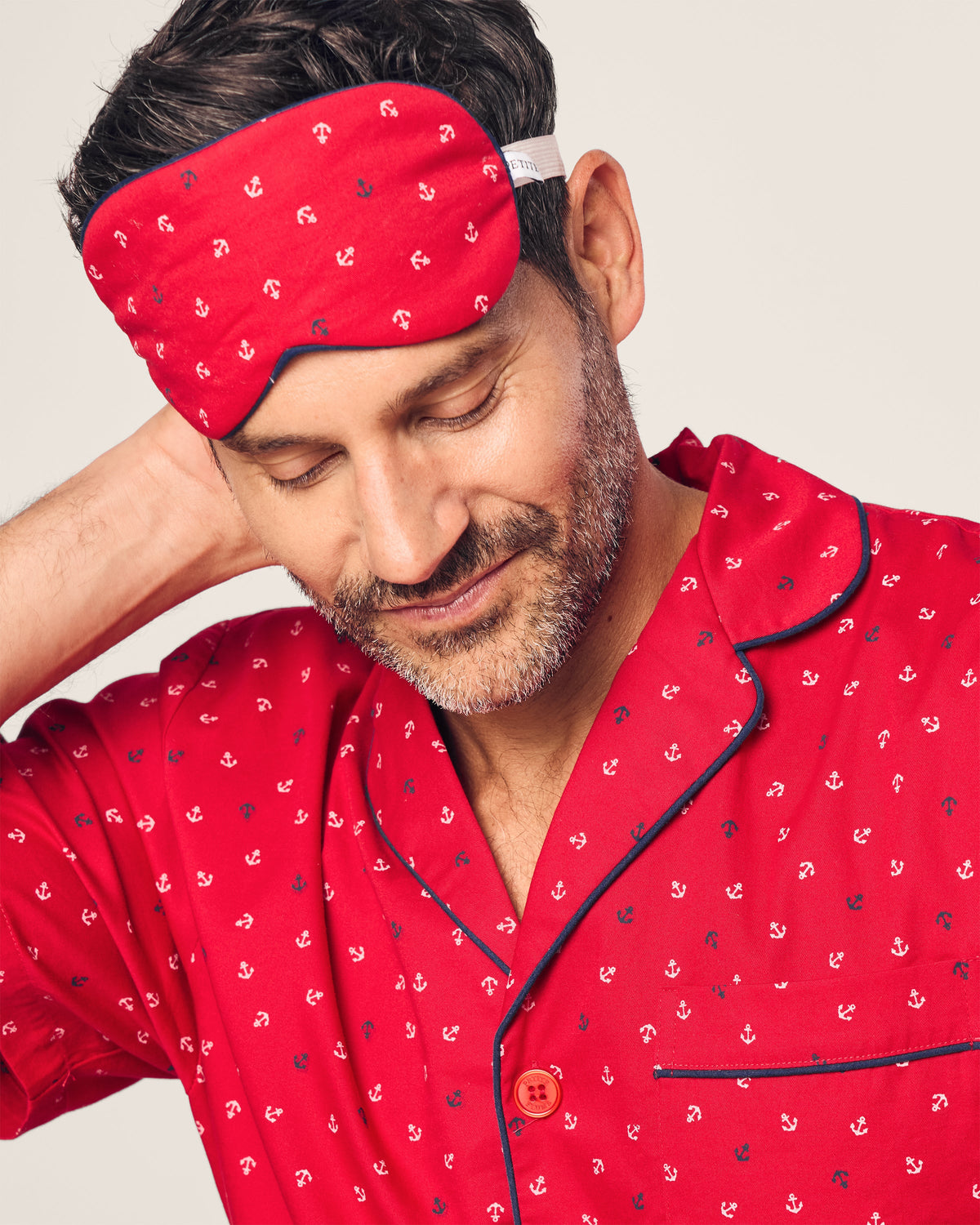 A person in red pajamas with a nautical print and wearing Petite Plumes Adults Twill Sleep Mask in Anchors Away scratches their neck, smiling contentedly. The background is a plain light color.