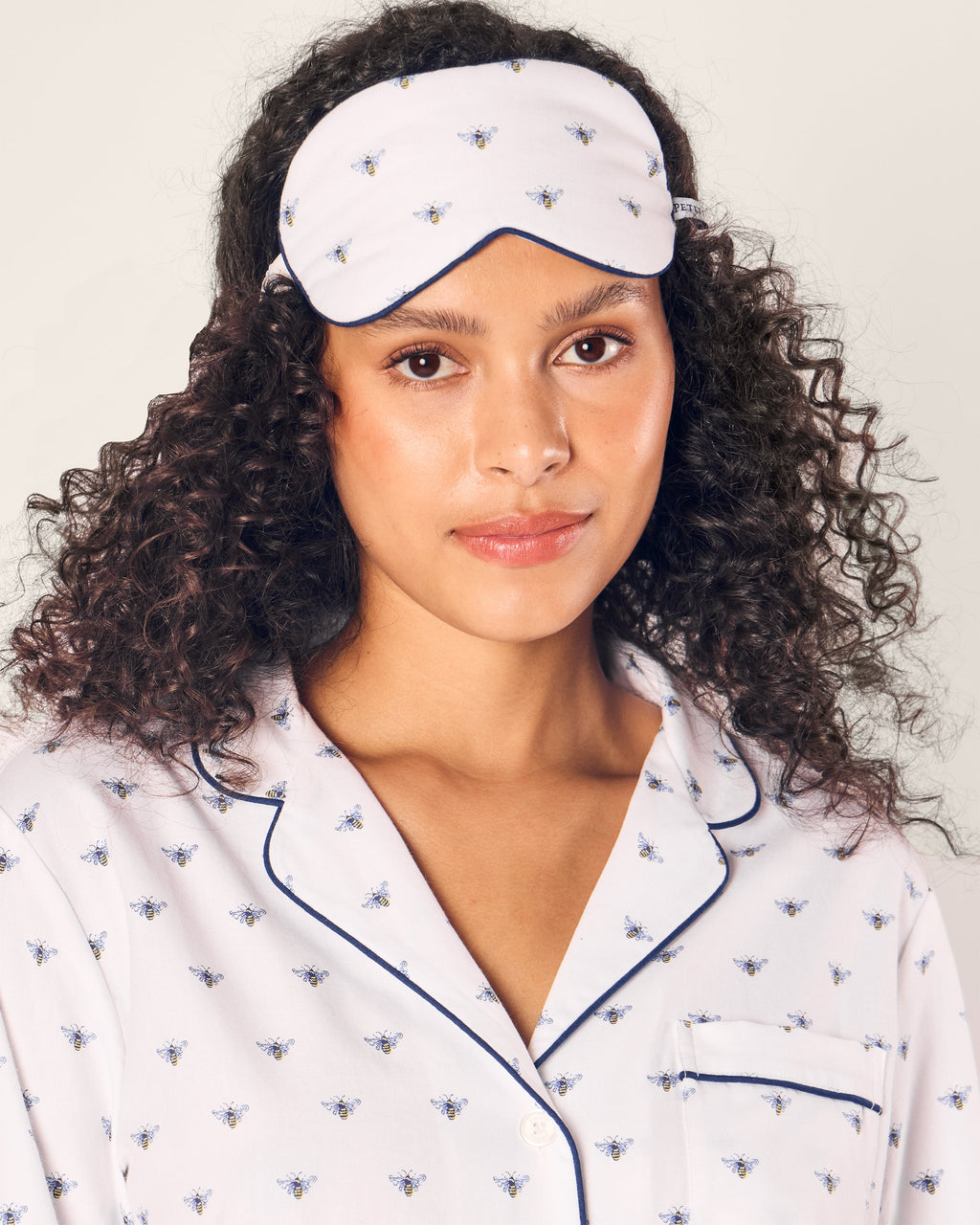 A person with curly brown hair wears Petite Plumes Adults Twill Sleep Mask in Buzzing Bees, paired with matching pajamas featuring charming bee patterns and dark piping on a plain, light background.