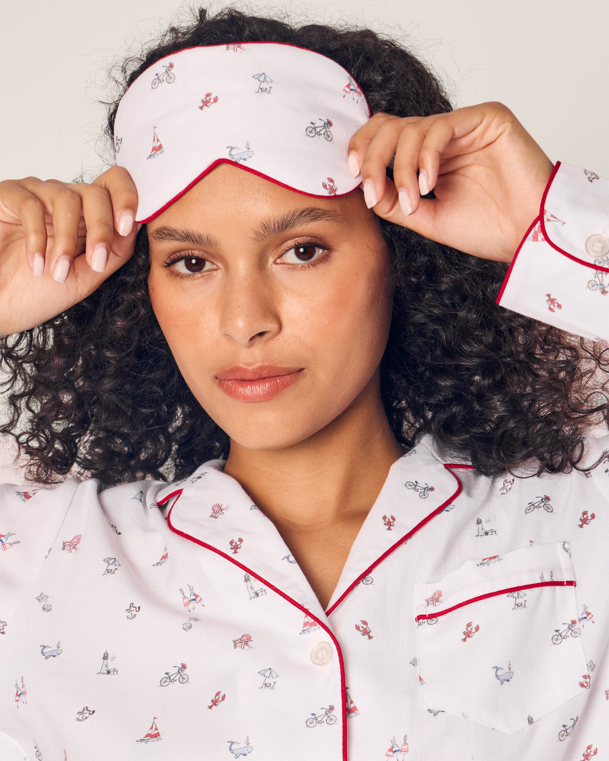 A person with curly hair adjusts an Adults Twill Sleep Mask in Coastal Charms by Petite Plume while posing in a white pajama top featuring bicycles and sailboats, looking directly at the camera.