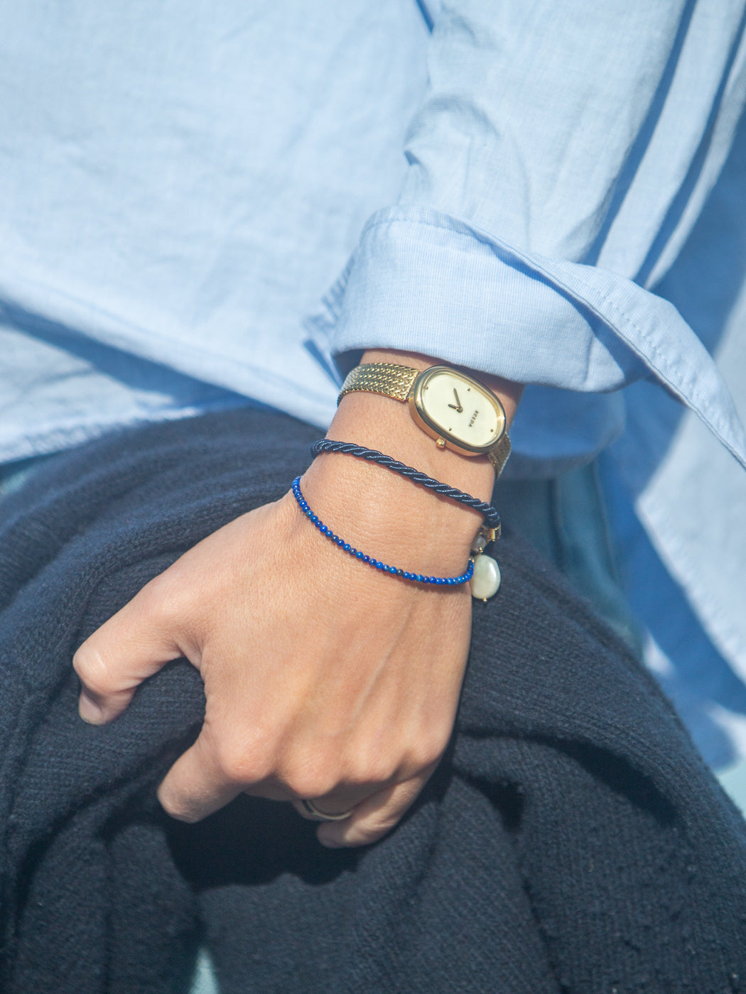 Model’s wrist wearing Alice bracelet in blue lapis beads with a freshwater pearl, layered with a gold watch and navy cord