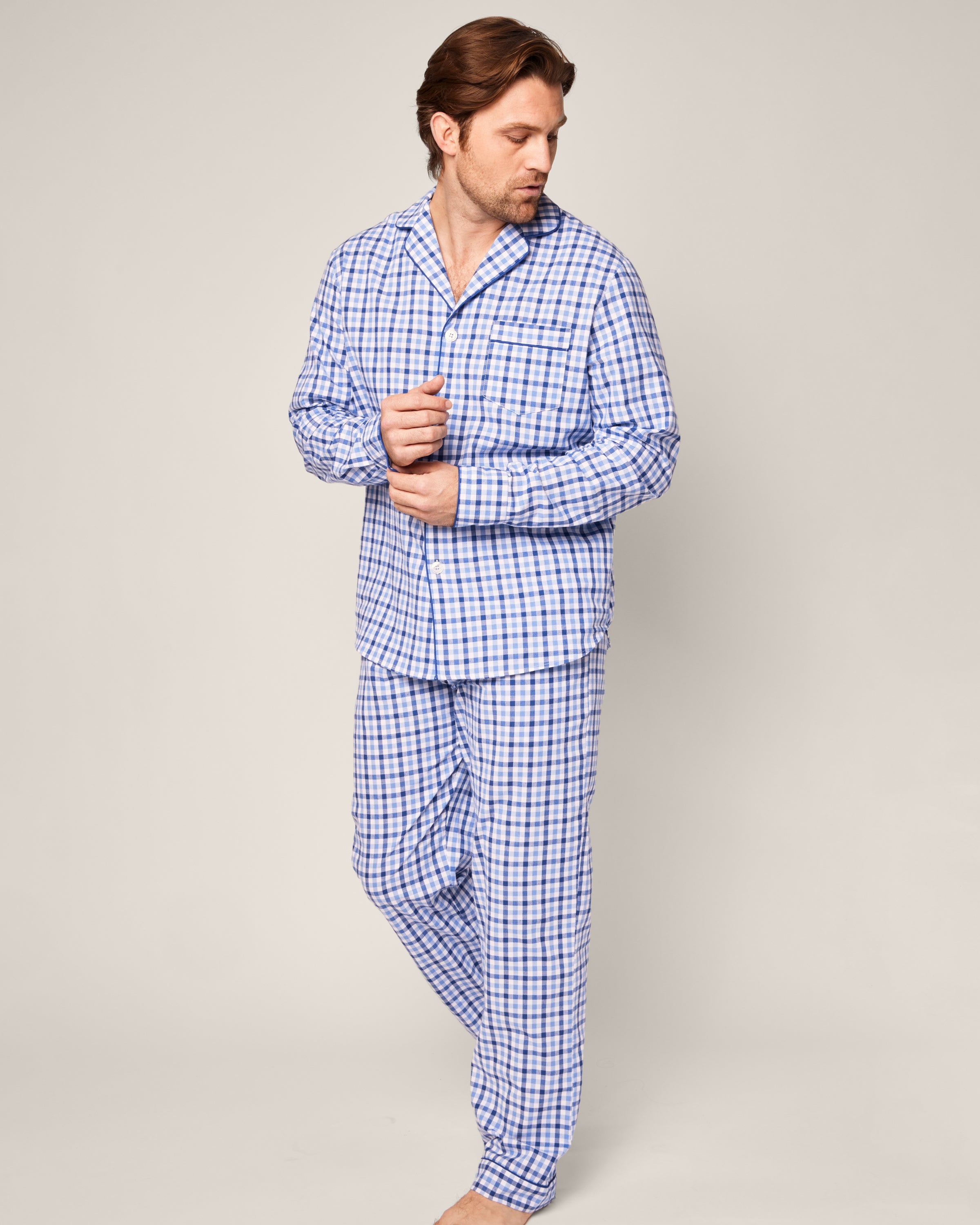 A man stands barefoot against a plain background, adjusting the sleeve of his Mens Twill Pajama Set in Royal Blue Gingham by Petite Plume.