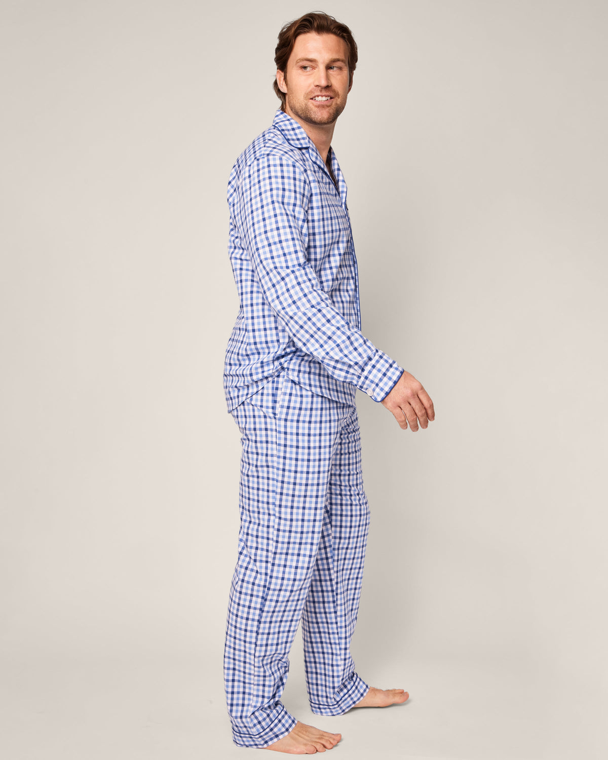 A person stands barefoot, turned slightly to the side, wearing the Mens Twill Pajama Set in Royal Blue Gingham by Petite Plume. They have short dark hair and smile against a neutral beige background.