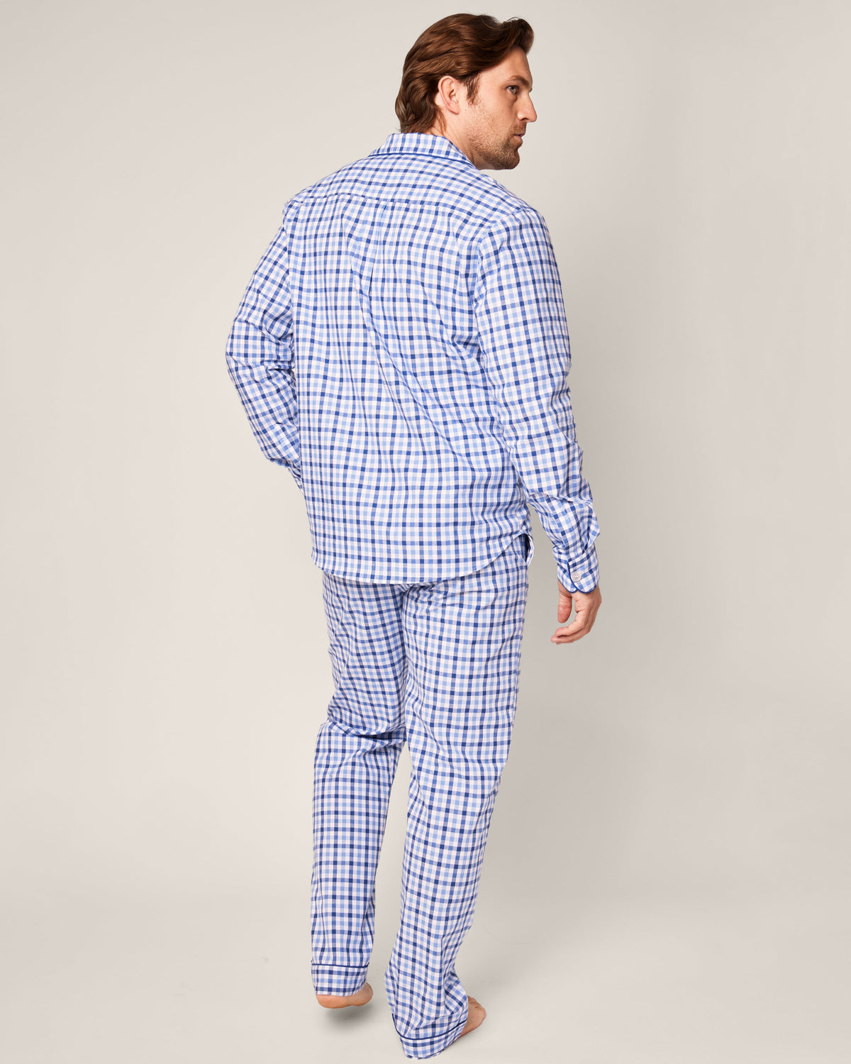 A man wearing Petite Plumes Mens Twill Pajama Set in Royal Blue Gingham, crafted from the finest quality cotton, stands barefoot on a plain background, slightly turning his head to the side.
